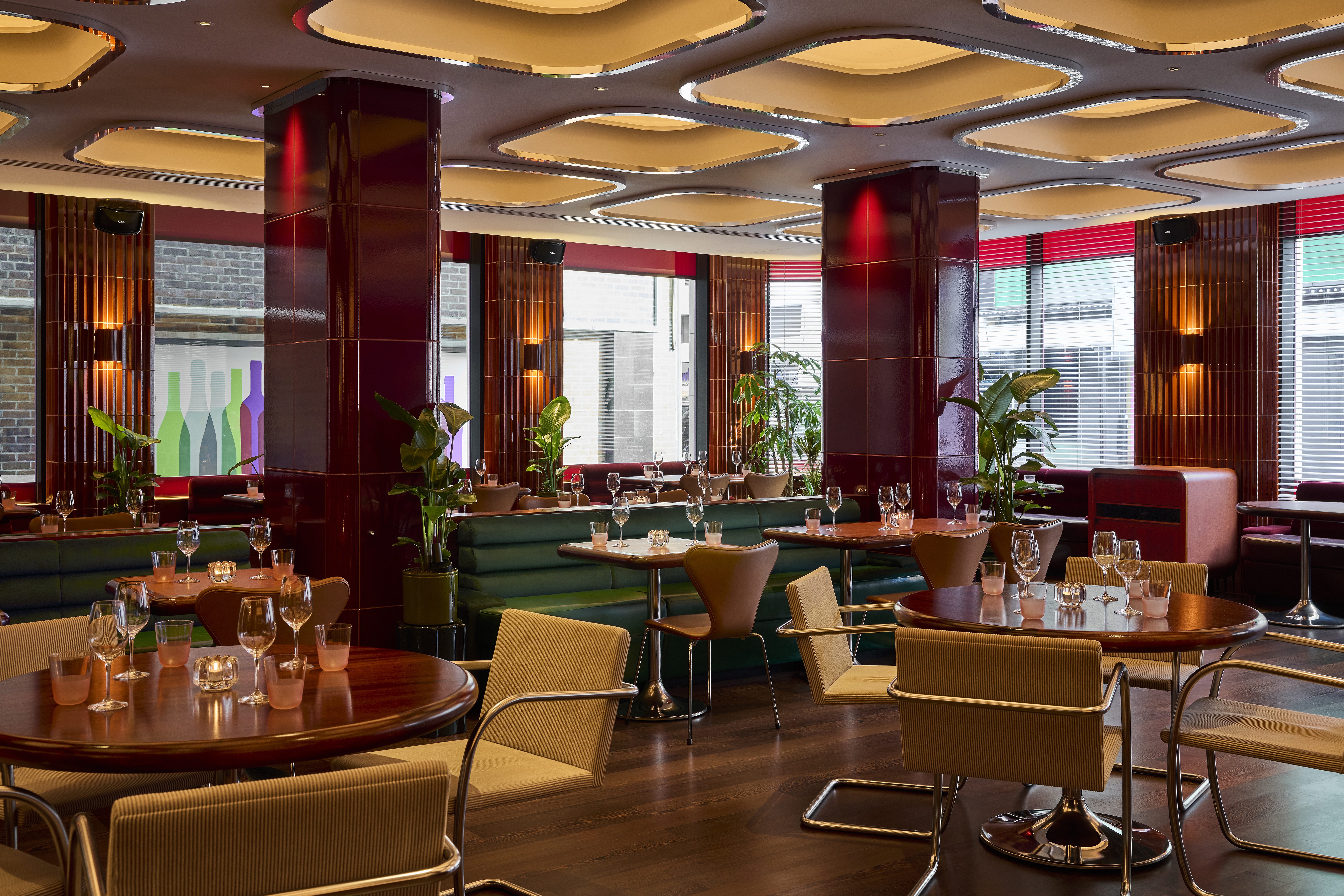 The interiors of a fine dining restaurant feature coffered, back-lit ceilings with chrome detailing, cherry plum walls covered in glazed ceramic, dark green banquettes, and atmospheric sconces, as well as plants.
