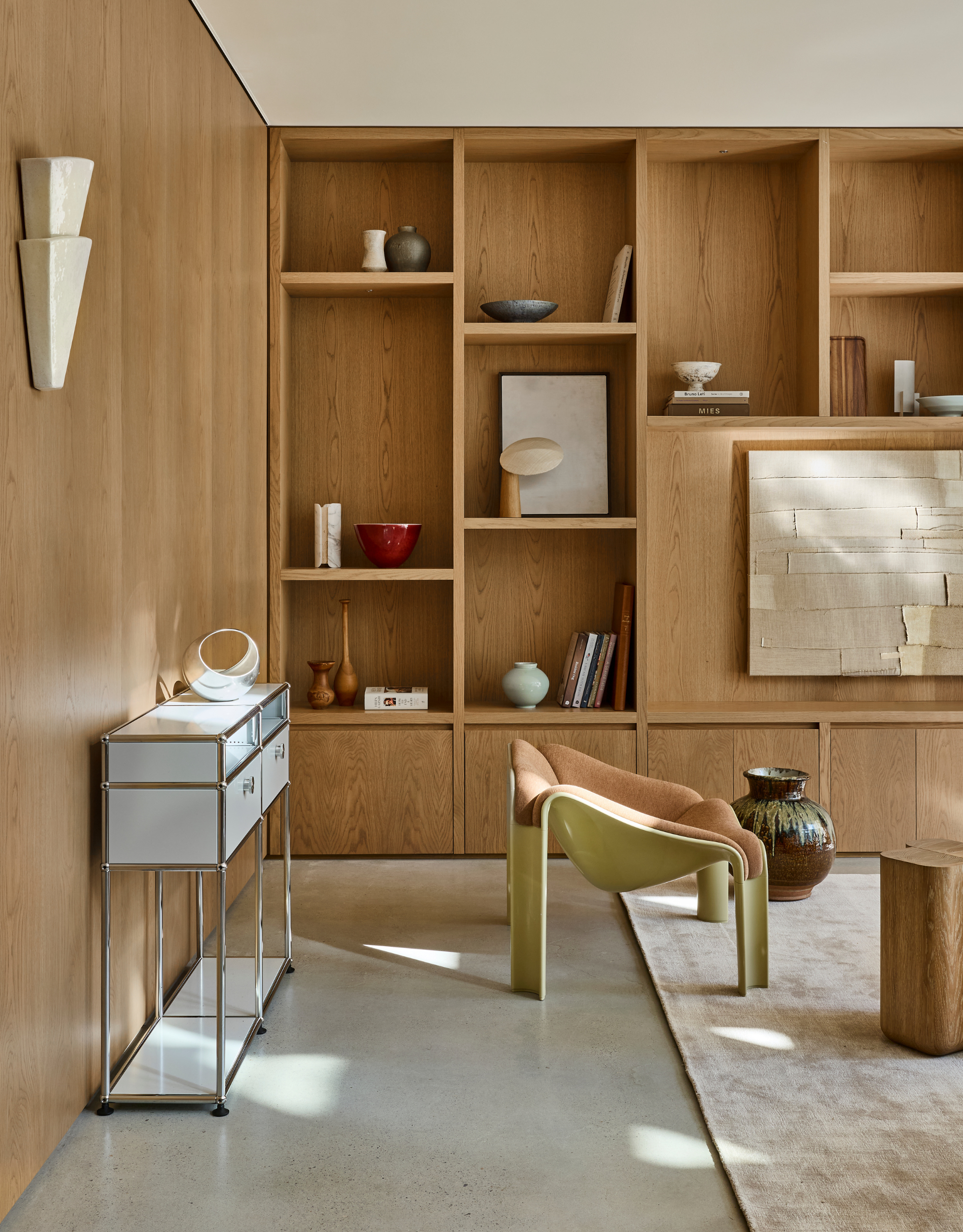 A bright minimalist living room with a gallery of ceramics and books, a chrome and white storage sideboard, a beige rug, a light brown accent chair, and a large vase