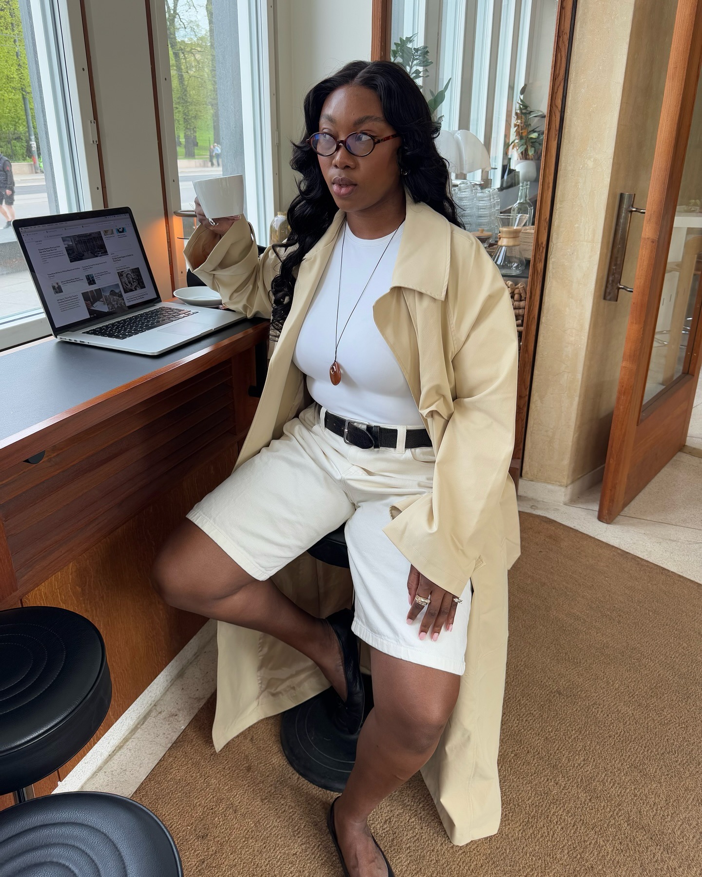 Influencer Nnenna wears a white denim Bermuda short outfit pairing them with a butter yellow trench coat a white fitted t-shirt and a black belt. She pairs this outfit with black ballet flats and a birthstone necklace in a photo on her instagram recently.