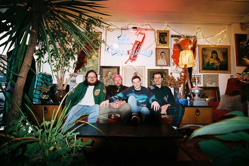 Image shows the four members of the band Pup sitting in a row. They are all white males. The wall behind them is decorated with pictures are there are two large houseplants visible.