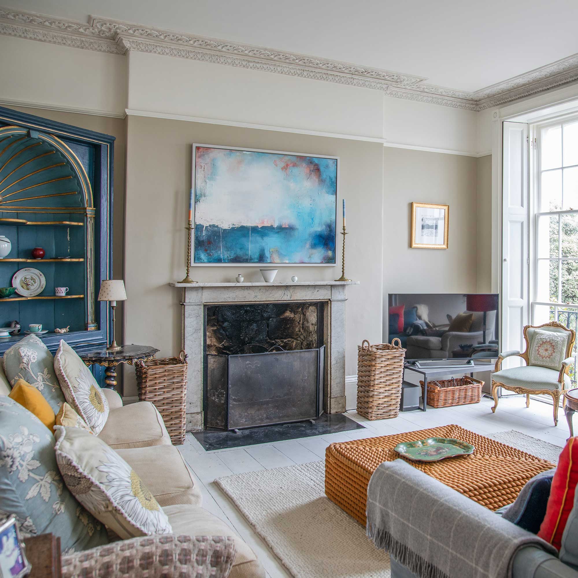 living room in a georgian villa with ornate coving, large sash windows, painted alcove cupboard and large landscape painting over fireplace