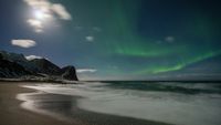 Aurora borealis lights up the night sky above a snowy mountain and beach shoreline, with waves gently washing onto the sand.