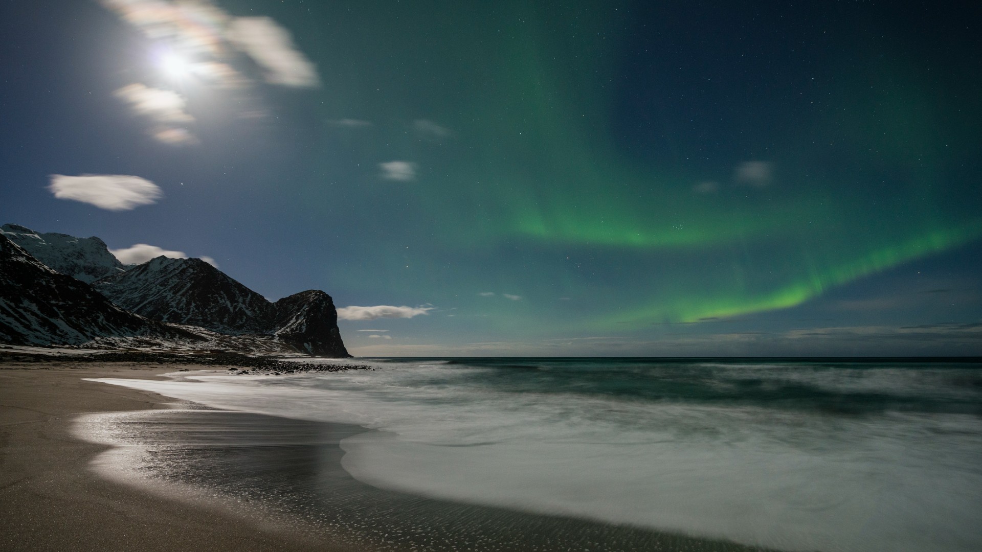 Aurora borealis lights up the night sky above a snowy mountain and beach shoreline, with waves gently washing onto the sand.