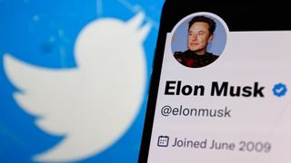The Twitter logo on the left in the background, with a phone in close-up on the right showing Elon Musk's Twitter profile