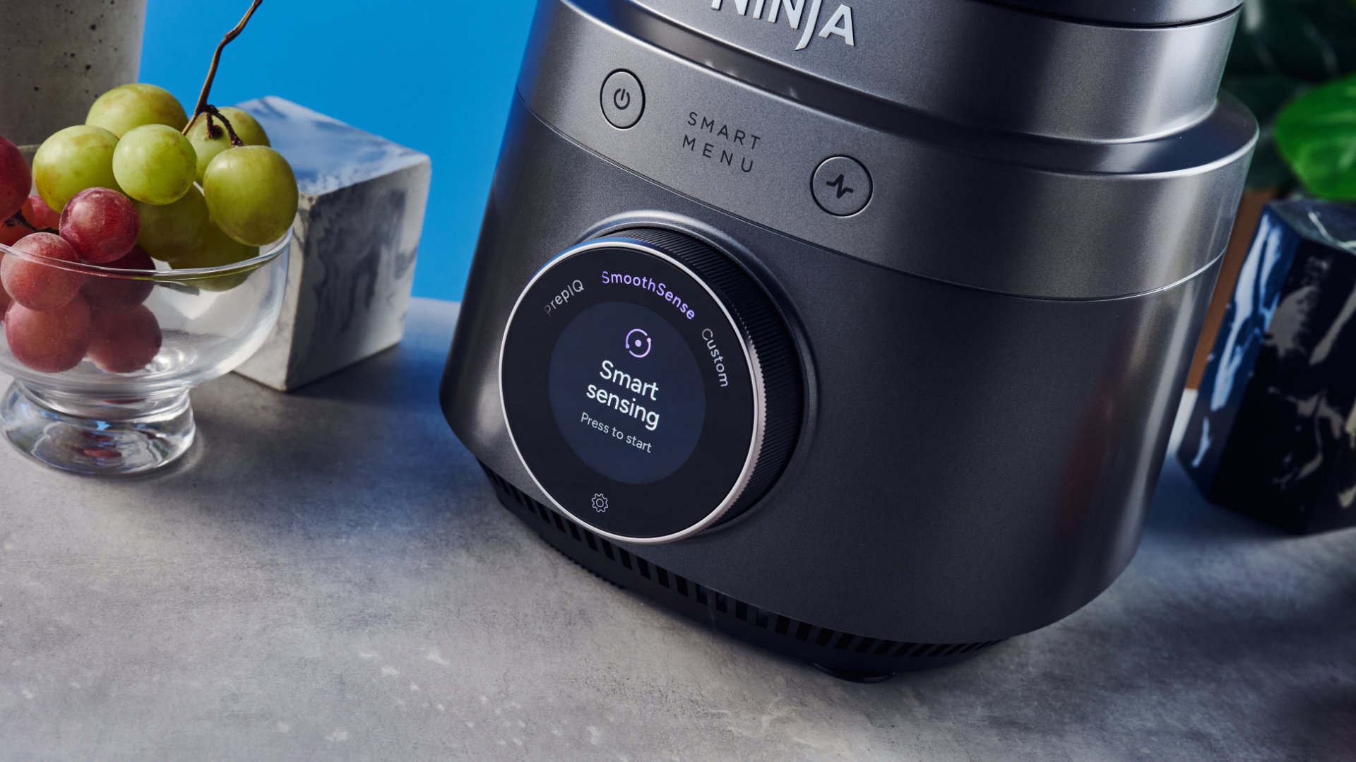 Ninja Crush Boss Blender in black photographed in front of a ble background with fruit.