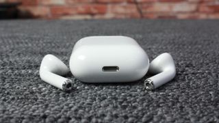 Ett par vita AirPods 2 ligger bredvid sitt tillhörande laddningsfodral på en grå textilyta.