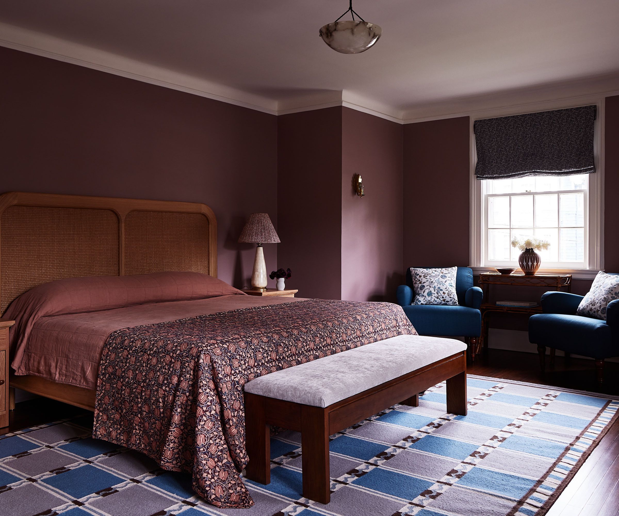 Burgundy bedroom with dark burgundy walls, rattan headboard, burgundy bedding, bench, purple and blue rug and window with two navy armchairs