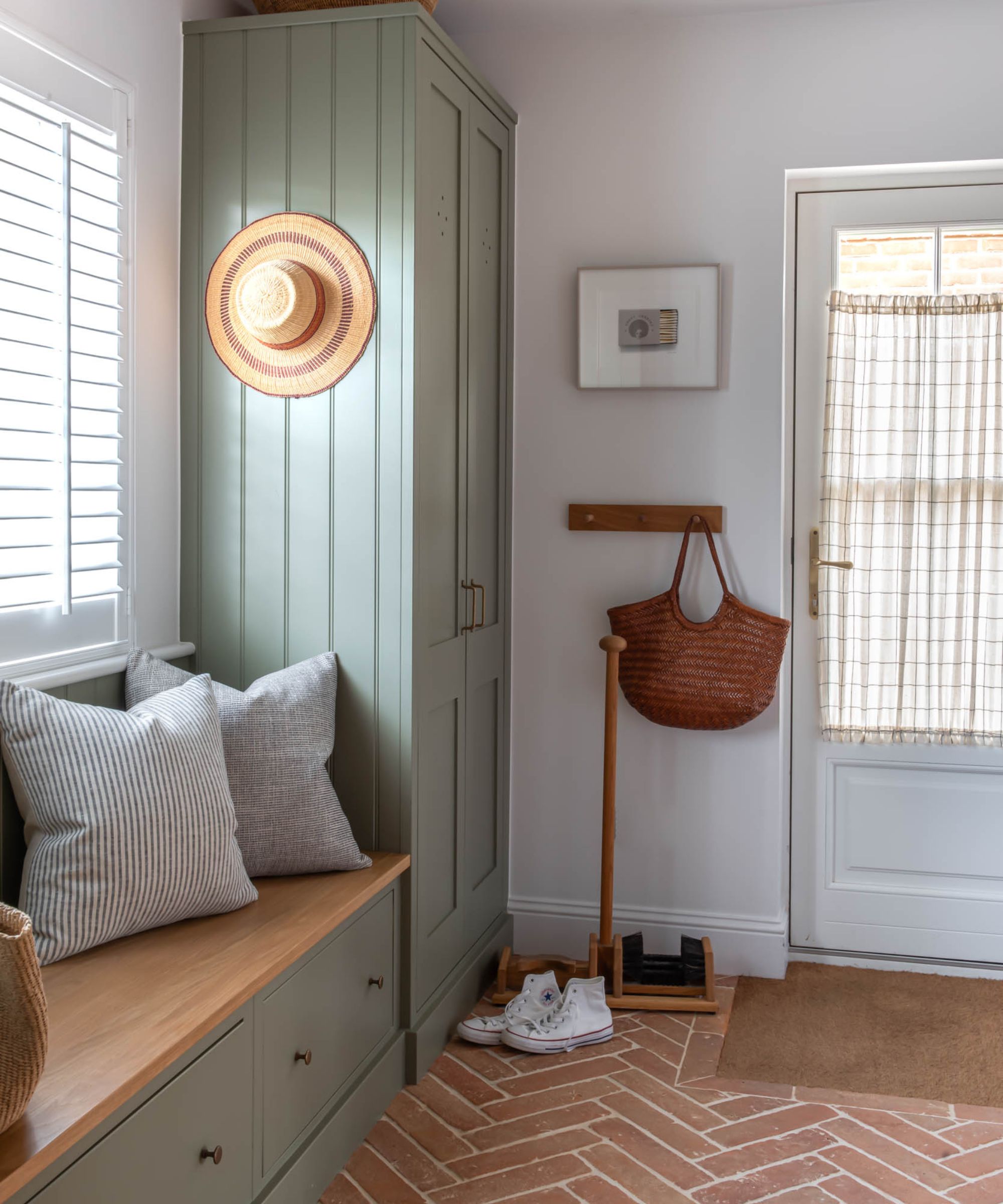 A mudroom with a built in bench and storage cabinet. Hooks beside the door, and a herringbone pattern brick floor.