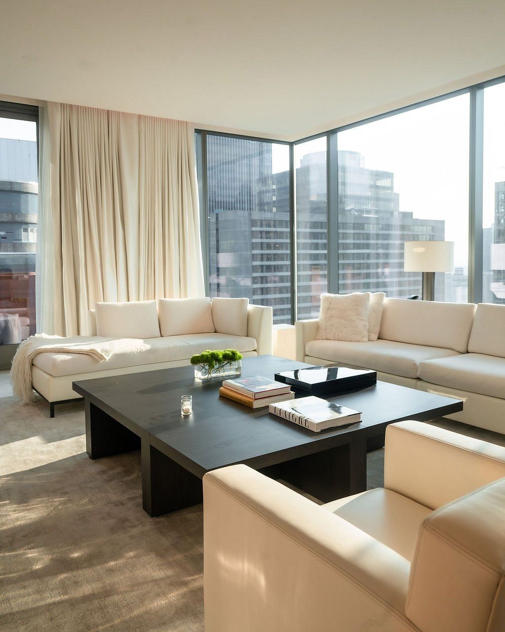 A superbly decorated, hyper-modern hotel features minimalist cozy furniture including neutrally-hued large couches, standing lamps, tiled floors, cream curtains, wall-to-wall windows, and breathtaking views of the city.