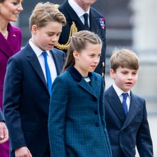 Prince George and Prince Louis wear navy suits with royal blue ties and white shirts, while their sister Princess Charlotte walks between them wearing a dark green and navy checked coat