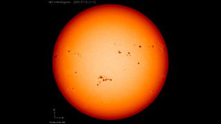 large orange red sphere which is the sun, against the black backdrop of space. Dark black patches are visible speckled across the surface of the sun, these are sunspots. 