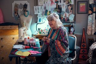 Susan Ruttan as Gladys, sitting in a room full of sewing supplies, a mannequin, a triangular set of drawers, a framed Playbill, and a pegboard full of sketches, in episode 105 of A 'Man on the Inside.'