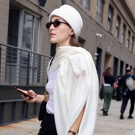 Editor Anna LaPlaca wears white pillbox hat, white scarf, black sunglasses and gold broach while walking on the street during a New York Fashion Week street style photo session.