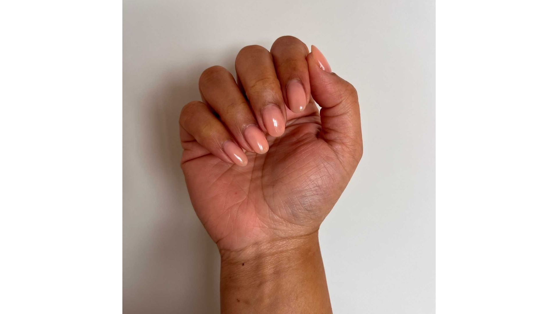 A close-up of Digital Beauty Ecomm Editor, Aleesha Badkar's hand, pictured with peachy sheer neutral nails, in front of a white wall background