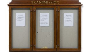 A wooden display case with three glass-fronted sections, each containing a white sheet of paper with text, and the word "TRANSMISSION" in gold letters across the top.