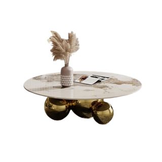 A round marble coffee table with three gold spherical legs, with a vase of pampas grass and an open magazine on top of it