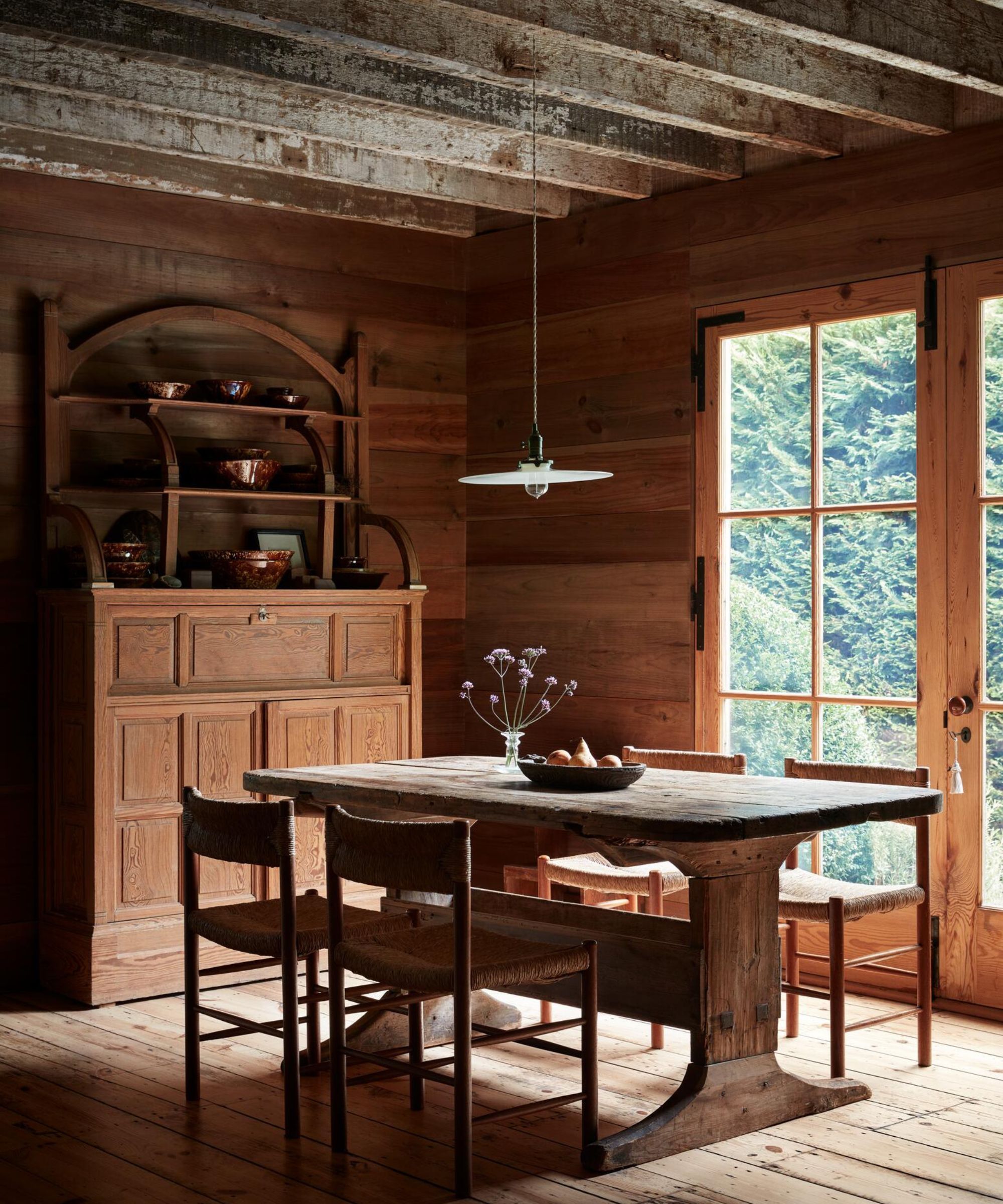 A rustic dining room with a heavy, primitive wooden table and four woven-seat chairs. Sunlight streams through a large paned glass door, highlighting the natural wood textures of the walls, ceiling beams, and a tall antique cabinet.