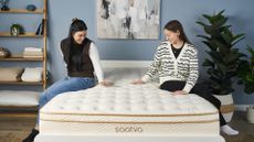 Two women sit on the Saatva Classic mattress in a bedroom. they're looking at the mattress trying to decide if this is the right one for their sleep needs