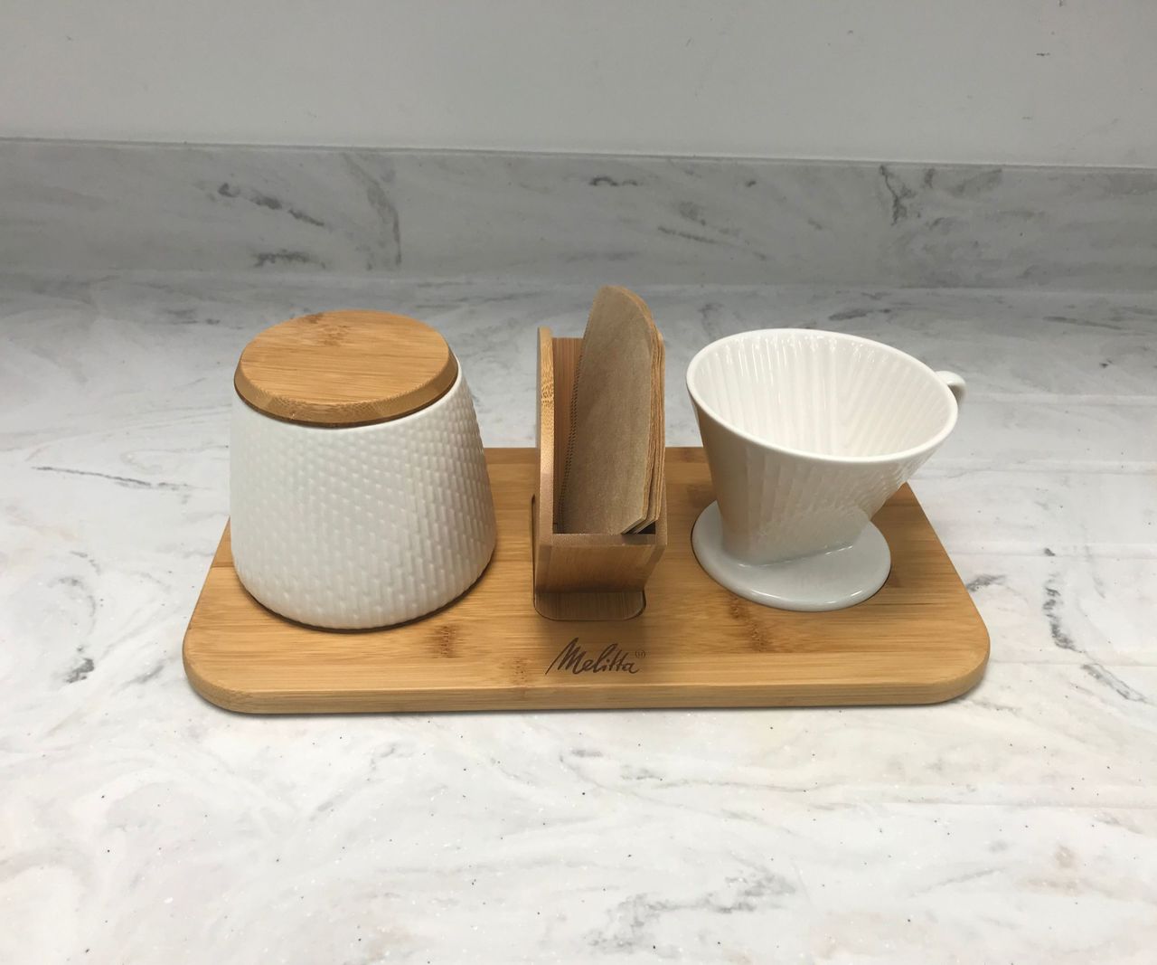 A wooden tray with a white pot and wooden lid, a container for holding filters, and a pour-over coffee maker on it.
