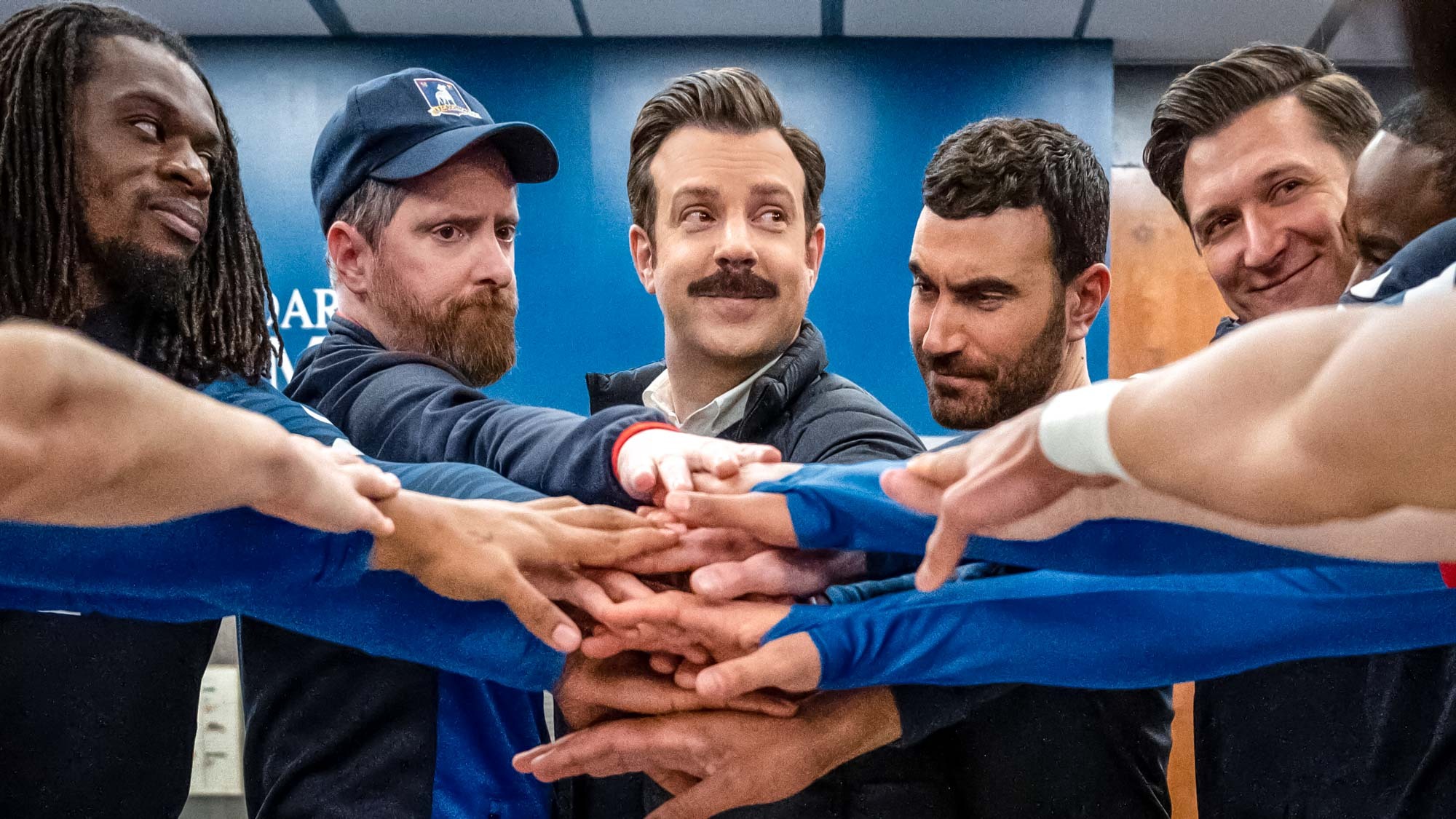 (Von links nach rechts) Brendan Hunt als Coach Beard, Jason Sudeikis als Ted Lasso und Brett Goldstein als Roy Kent, der vor einem Spiel in Ted Lasso Staffel 3, Folge 12, seine Hände in die Mitte legt. 