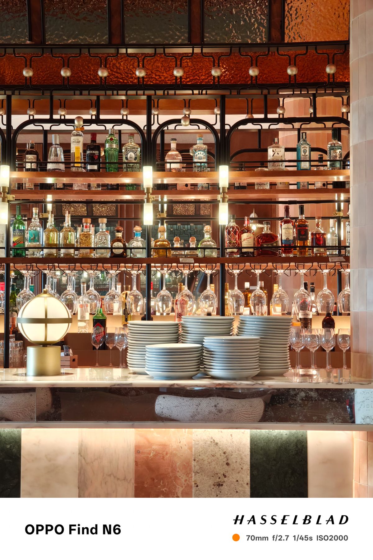A sophisticated indoor shot of a high-end bar at night. The image shows several rows of backlit liquor bottles on glass shelves, with rows of upside-down wine glasses hanging below. In the foreground, neat stacks of white ceramic plates sit on a marble countertop next to a spherical, brass-based lamp.