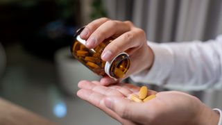 Close-up image of a person pouring some yellow pills from a semi-transparent bottle onto their hand. They appear to be wearing a white shirt. The bottle of pills has a white plastic rim.