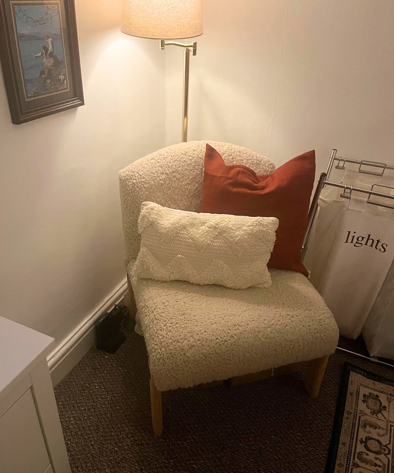 A teddy fabric chair in the corner of a bedroom, two pillows on it, a swing arm lamp behind, and a separated laundry hamper next to it.