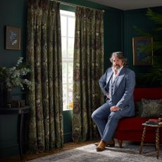 Terry and House Llewelyn-Bowen fabric curtains in dark green in a green room with man sitting next to them on a sofa.