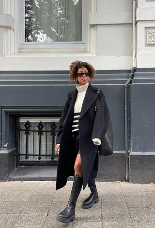 how to wear Chelsea boots is shown in a photo of a woman standing outside wearing a long black coat over a black and white stripe turtleneck sweater styled with black shorts, a black hobo tote bag, and black knee-high chelsea boots