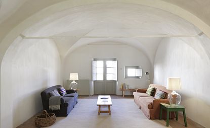 A room in the São Lourenço do Barrocal hotel. White walls with domed ceiling, light gray doors, and window, deep blue couch to the left, orange couch to the right with a wooden table in between them.