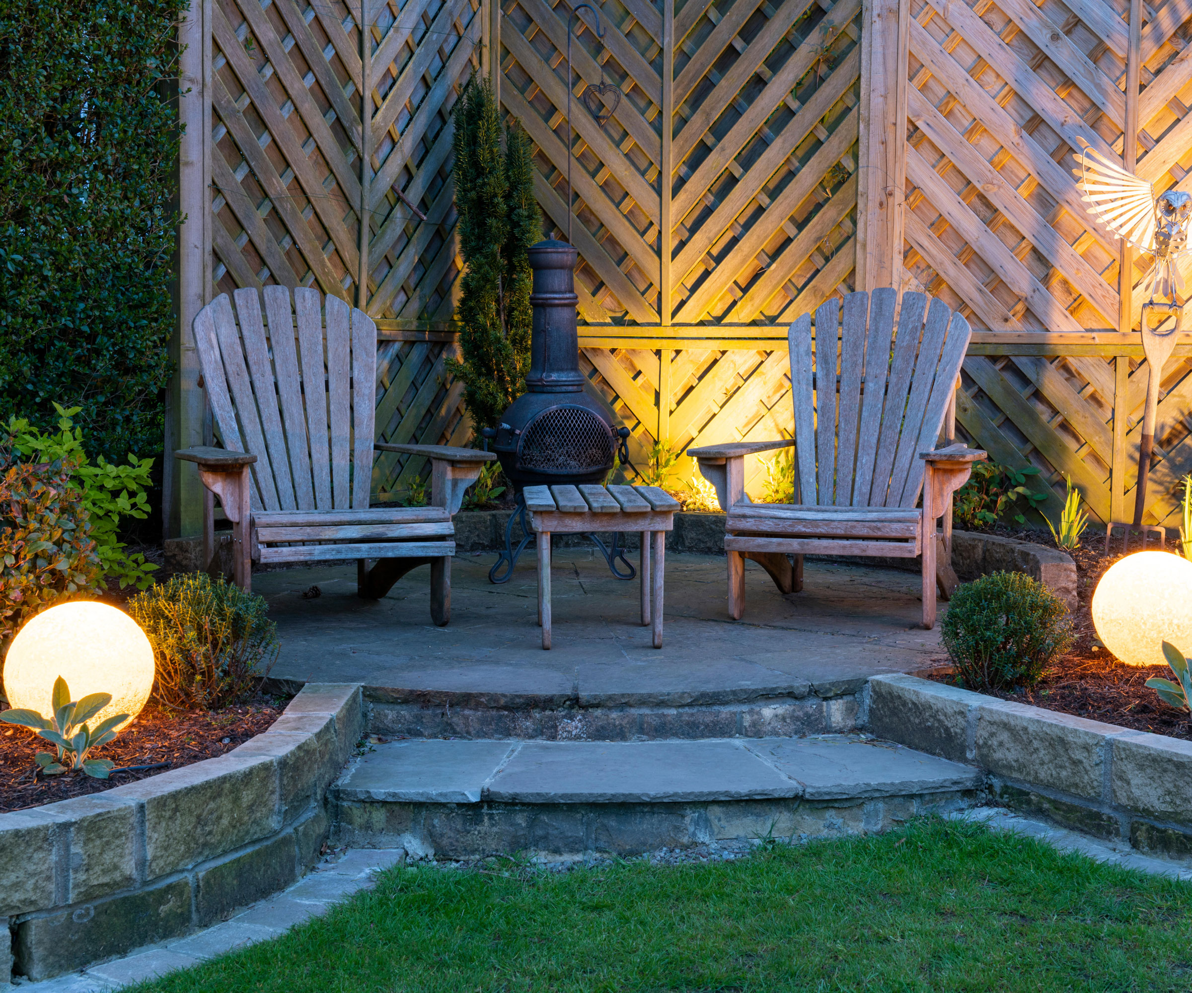 pair of Adirondack chairs with small table, chiminea, trellised fence on raised patio area, with pair of white glass globe lights illuminating raised flower beds
