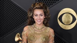 Miley Cyrus is smiling and wearing a dress made entirely of gold safety pins at the Grammy's red carpet.