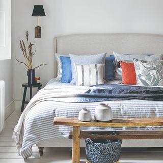 Double sized bed with striped grey bedding and coloured cushions, with bedside table and wooden table in front