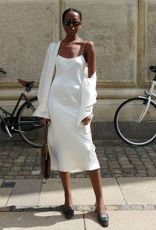 a woman's outfit showing what to wear in 80-degree weather with a white slip dress styled with a white button-down shirt, black mules, and a frame handbag