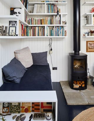 A garden studio living room corner idea with wood burning stove, open shelving with books and corner bench with blue and cream cushions