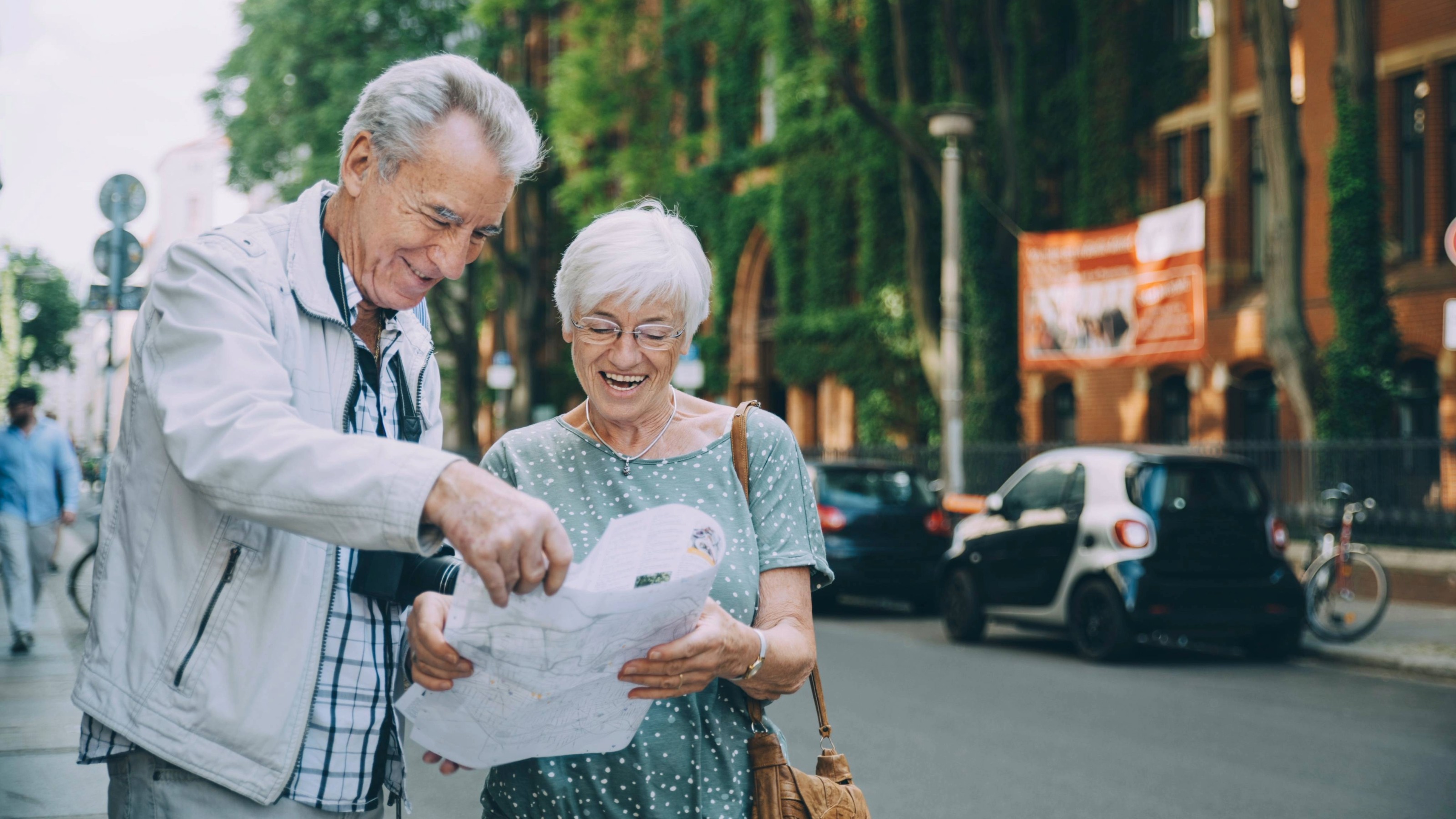 Smiling elderly couple reading map while standing on sidewalk in city during vacation