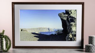 The Netgear Meural Canvas II showing a photo of rocky cliffs and an azure sea. It's surrounded by a plant and some patterned candle holders.