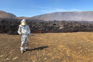 A man in a protective suit stands holding a long lava penetrometer