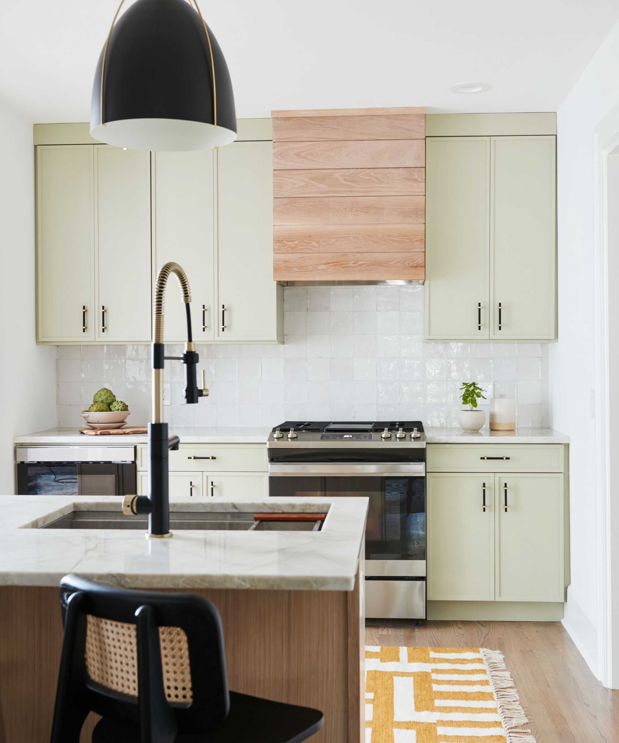 A modern, small kitchen with pale green cabinets, white wall tiles, wooden flooring with a yellow and white patterned rug, and a kitchen island with a stone countertop.