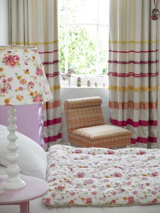 girls bedroom with floral quilt and lampshade, orange print chair, stripe curtains with fringing, pink side table, windowsill with Russian dolls