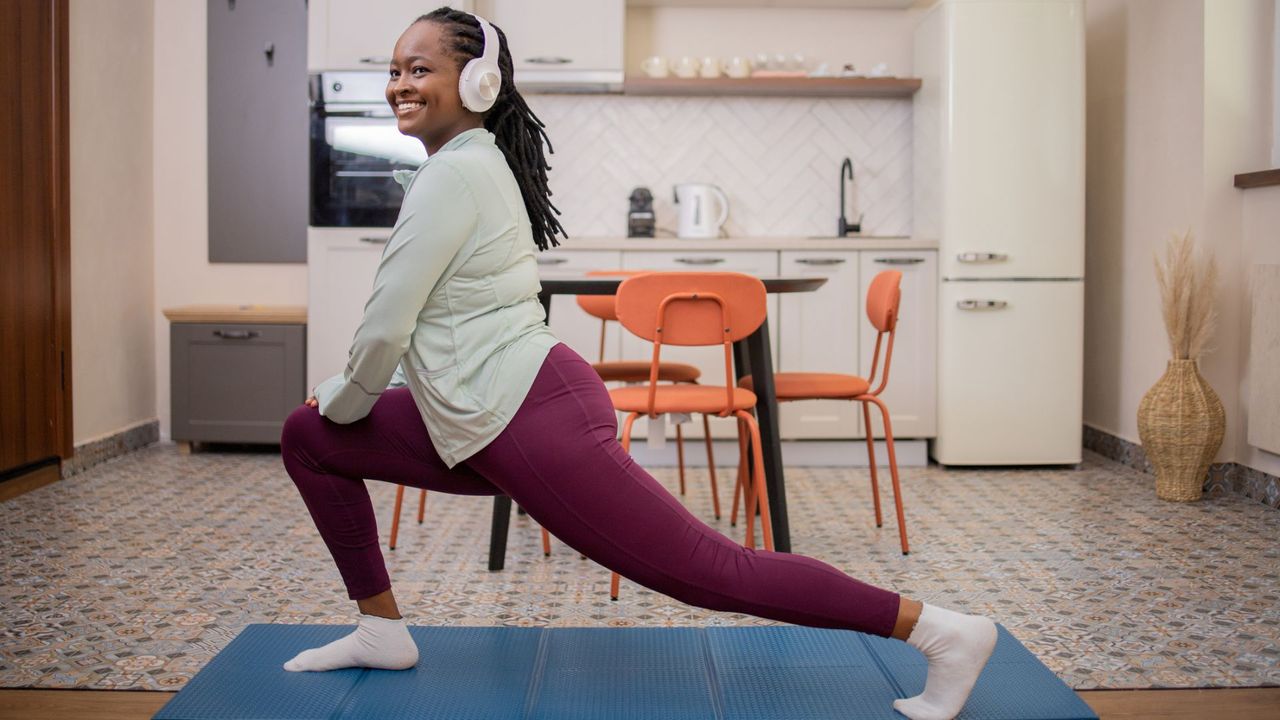 woman in headphones does a lunge stretch on a mat in her kitchen