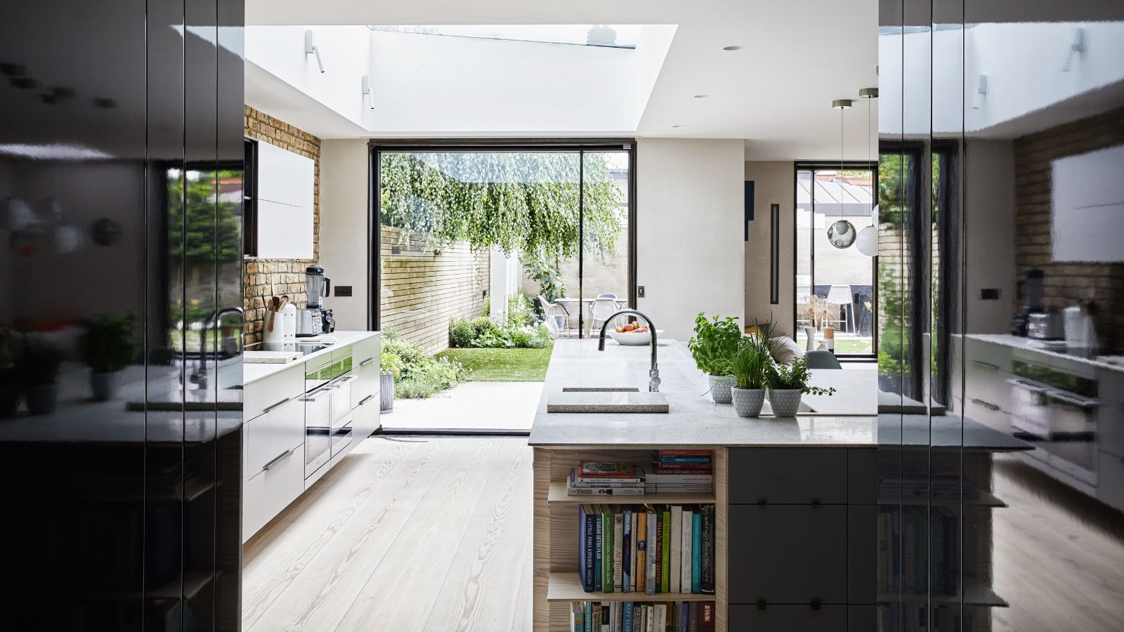 A bright and airy modern kitchen with wooden flooring, black and white cabinetry, an island with white marble countertops and built-in shelving filled with recipe books, an exposed brick backsplash, and large window doors to a lush green garden.