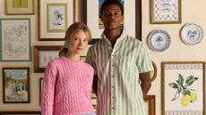A female model wearing a pink jumper from Joules, along with a male model wearing a striped green and white shirt from Joules, standing in front of a colourful photograph gallery wall.