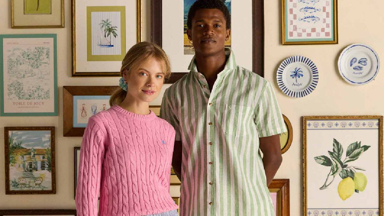A female model wearing a pink jumper from Joules, along with a male model wearing a striped green and white shirt from Joules, standing in front of a colourful photograph gallery wall. 