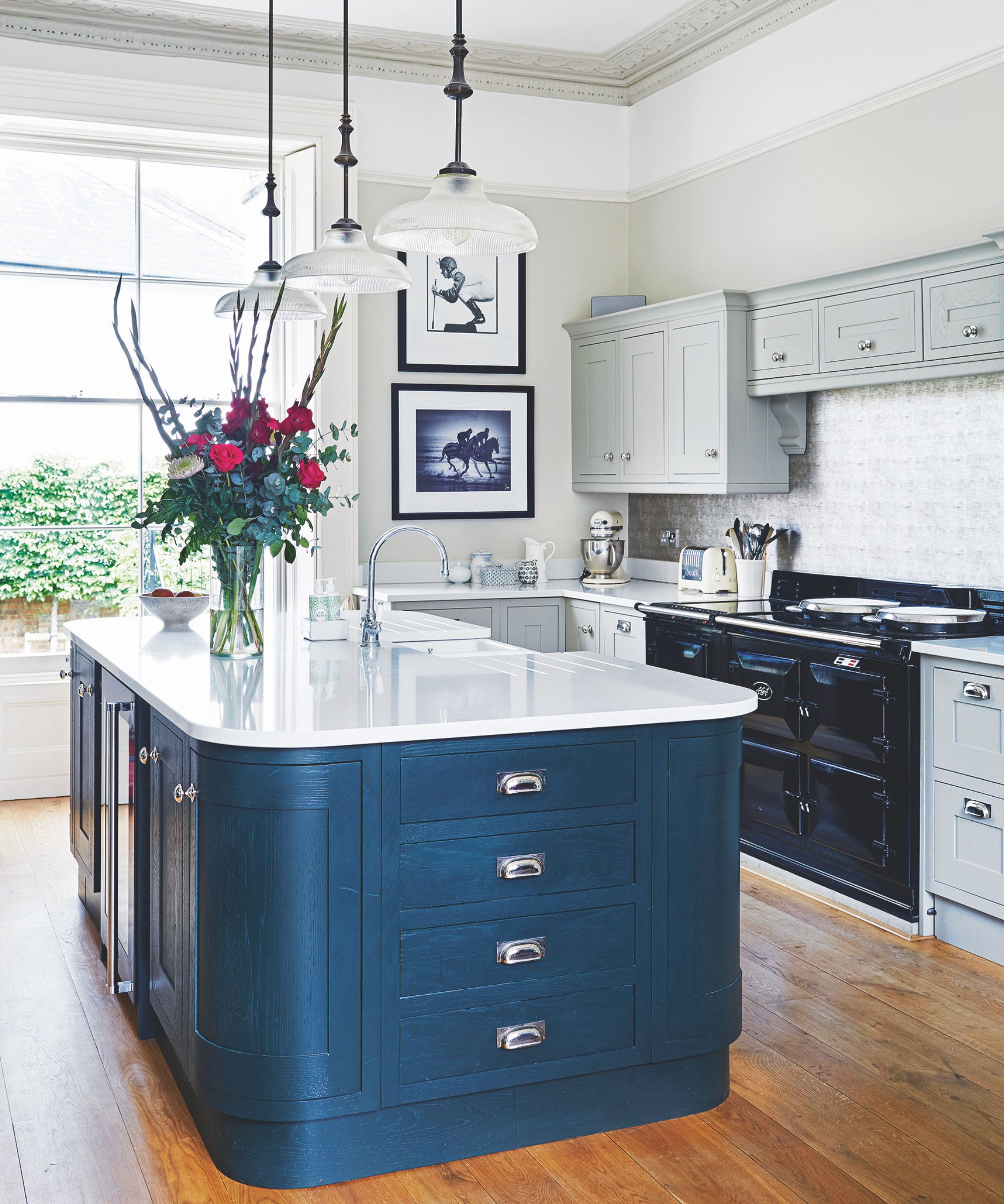 A white and blue kitchen with white countertops, wooden flooring, a black AGA and pale blue cabinets. On the island is a large bouquet of red flowers.