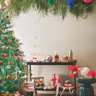 Christmas tree with brightly coloured streamers and baubles. Next to it is a side table with a champagne tower and presents.