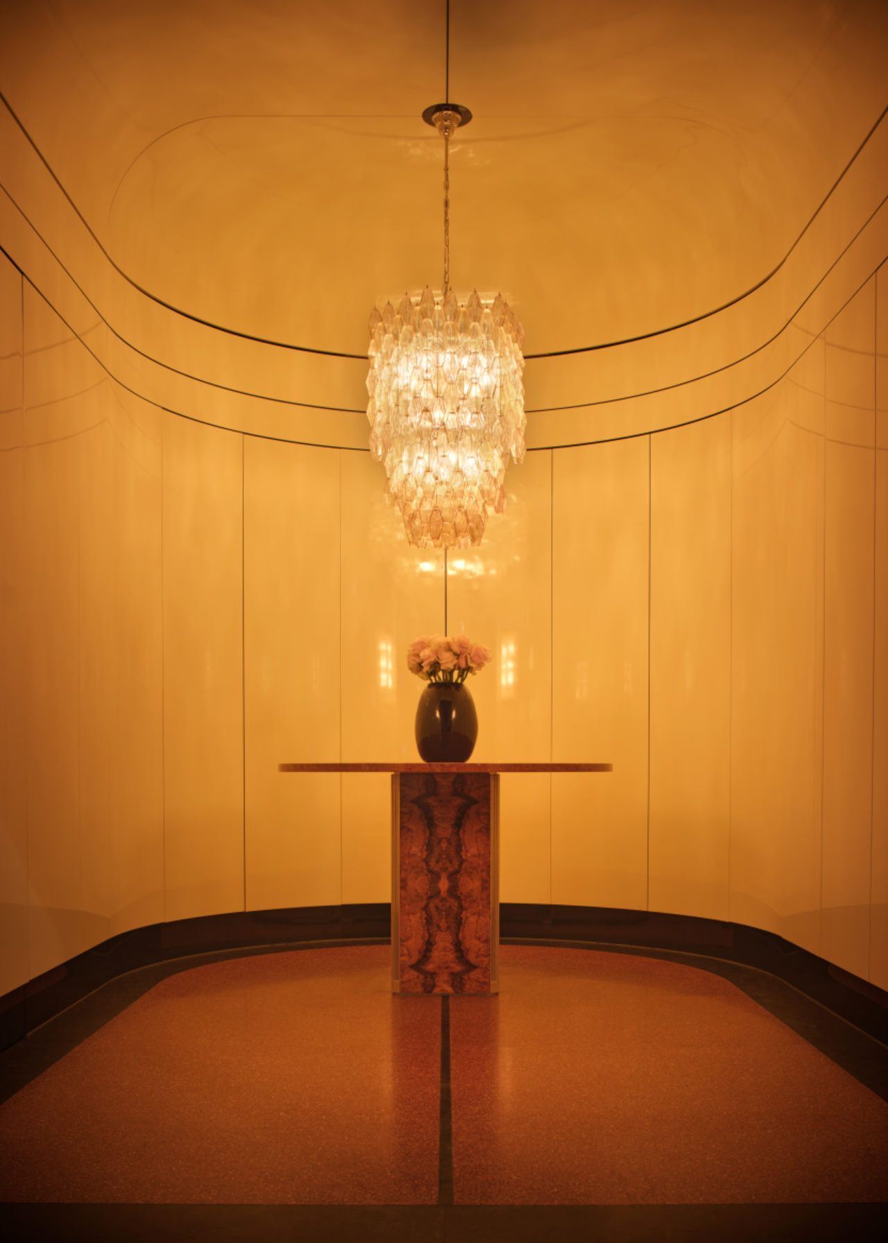 a curved room with white, glossy walls, and a large chandelier in the centre, hanging over a small wooden table