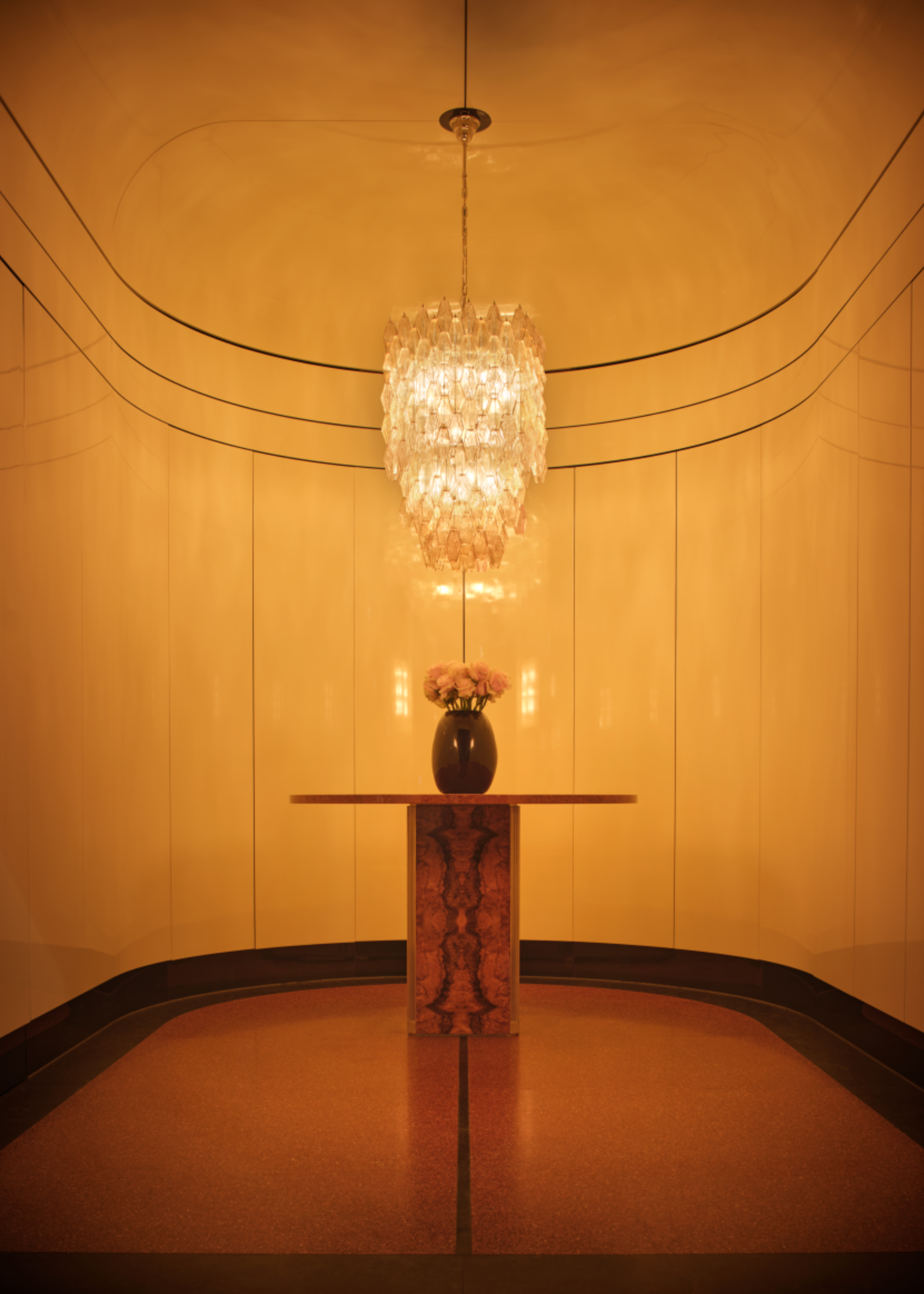 a curved room with white, glossy walls, and a large chandelier in the centre, hanging over a small wooden table