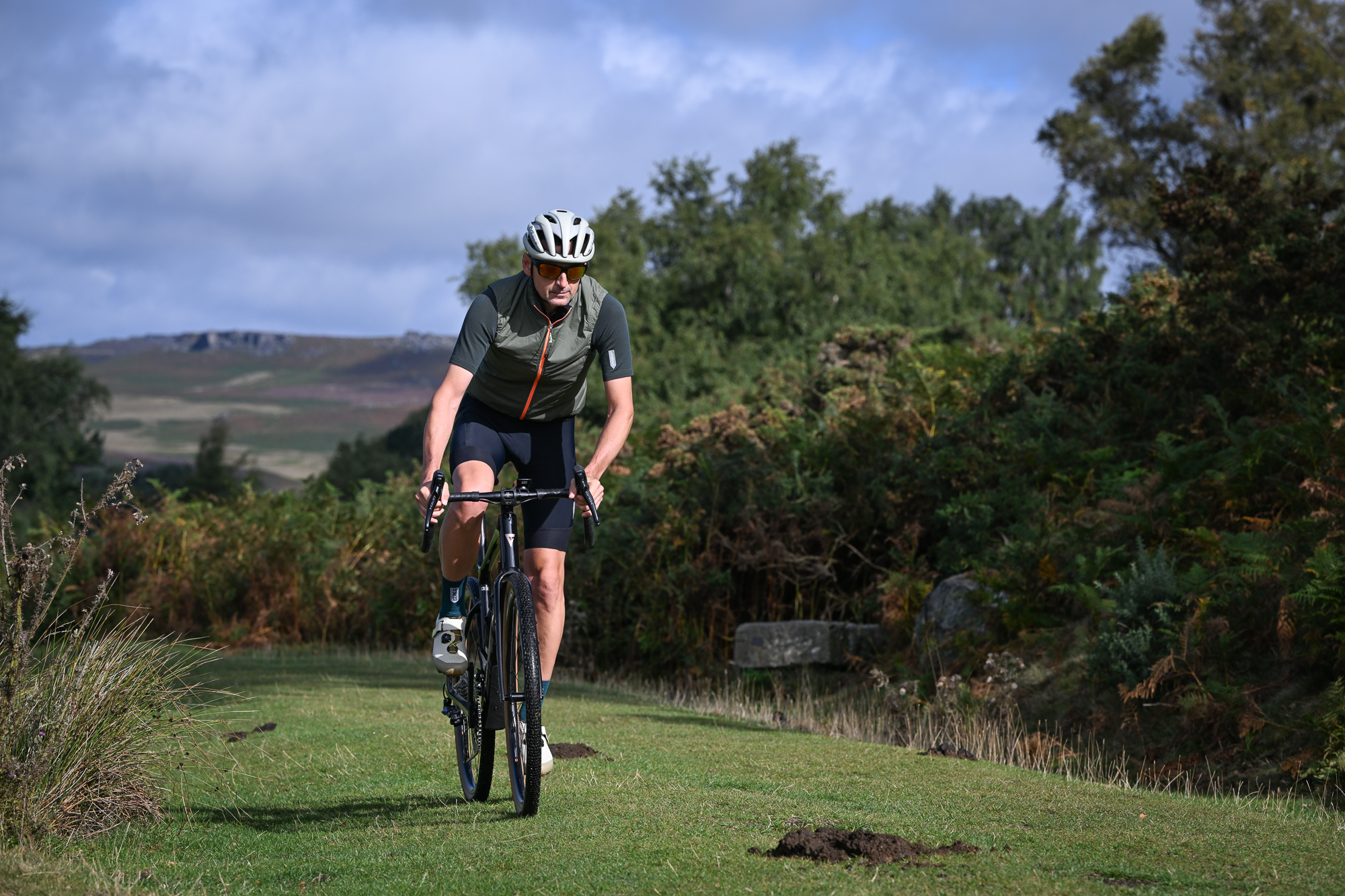 Man wearing a green gilet and dark green jersey riding a black gravel bike towards the camera on a grassy trail