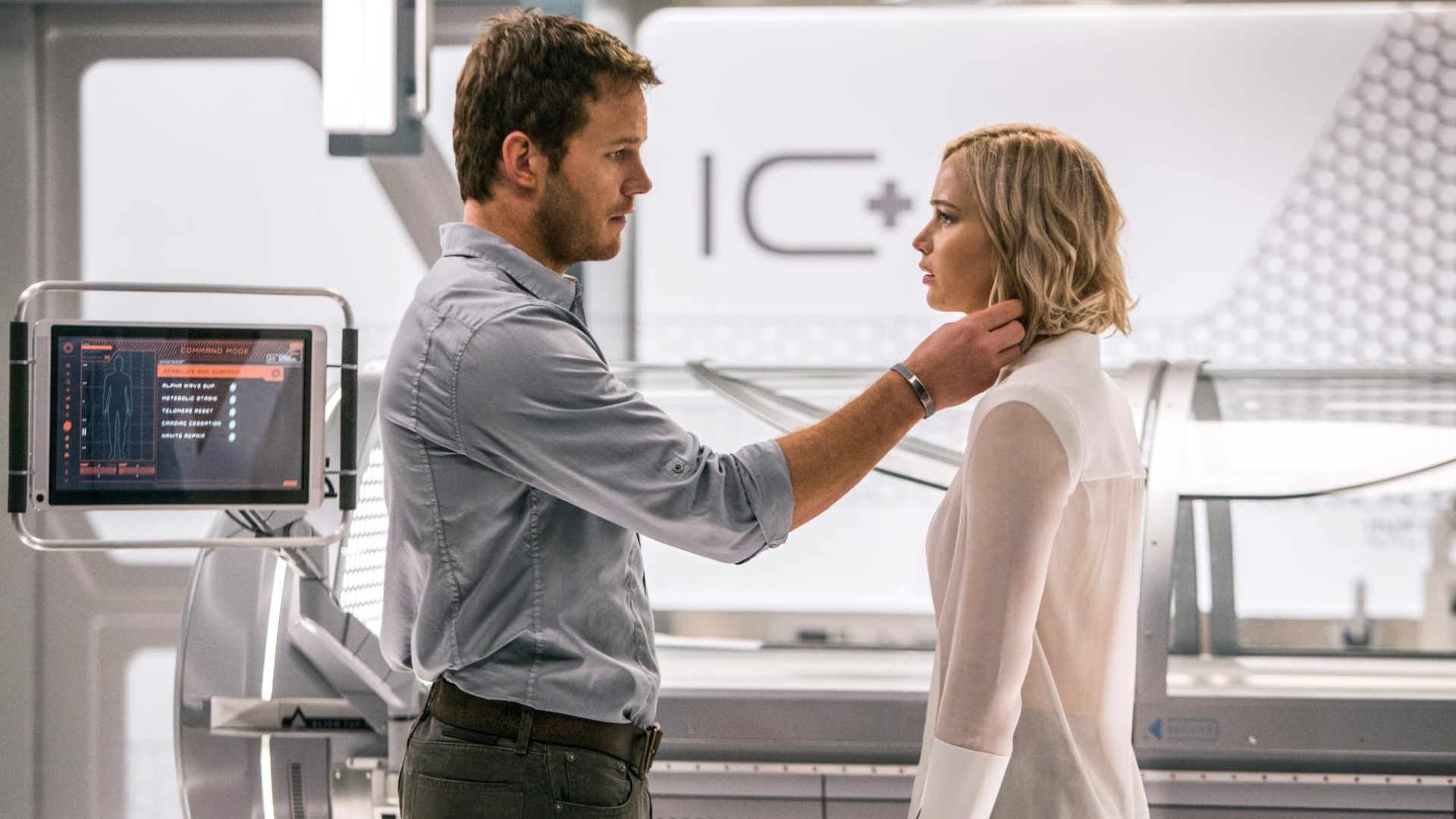 A bearded brown-haired man and a blonde woman in a sci-fi medical lab. The man is touching the woman's hair as they stare at each.